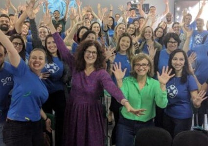 Specialist Dorothy Zemach in a purple dress, standing, right arm raised, left arm lowered, with a group of participants in blue shirts, arms extended
