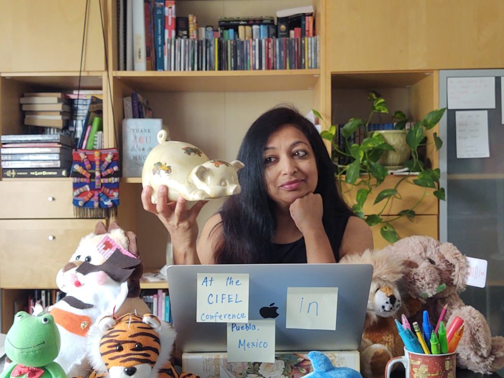 Specialist Natasha Agrawal holding a porcelain pig, left fist under her chin, in a seated position in front of a laptop