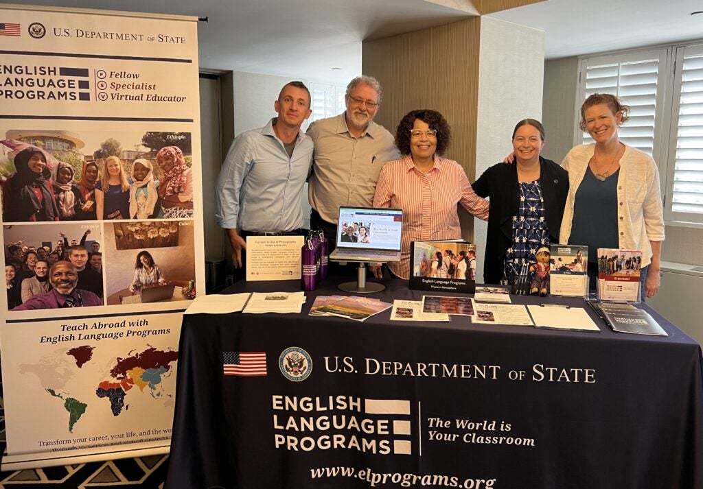 5 English Language Programs alumni standing behind an English Language Programs recruitment table, which is adorned with promotional materials
