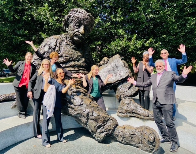8 English Language Programs alumni festively pose with their hands in the air around a statue of Albert Einstein