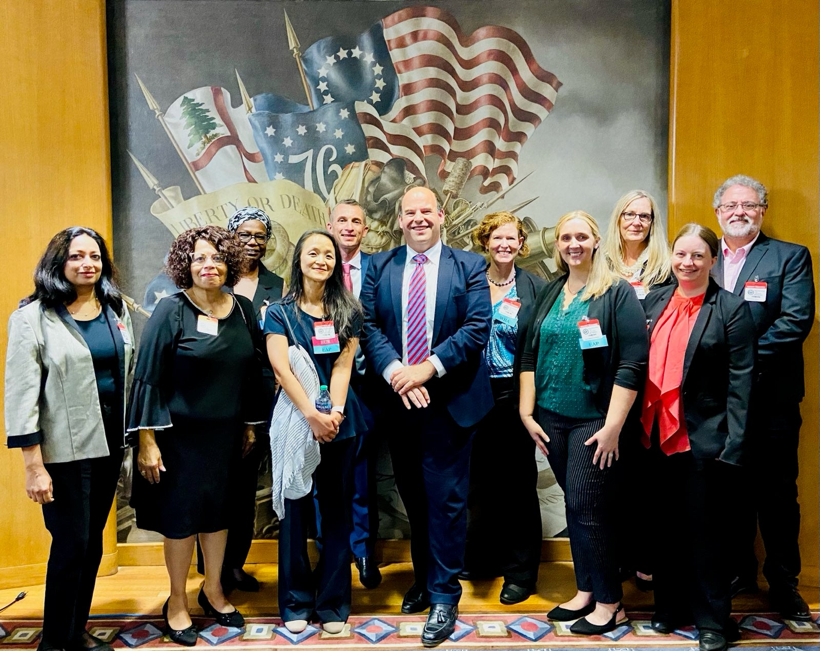 10 English Language Programs alumni with Deputy Assistant Secretary Ethan Rosenzweig pose in front of a large backdrop picture of U.S. flags