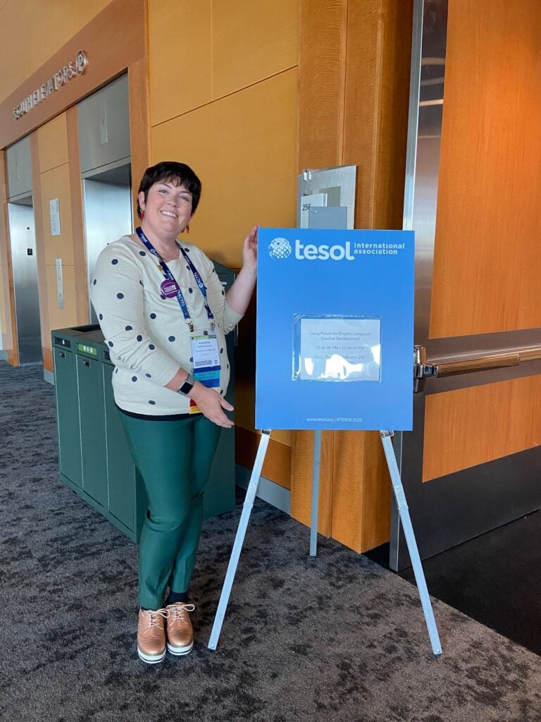 Katherine Anderson standing outside of a room posing next to a TESOL International Association convention session sign that is on an easel stand