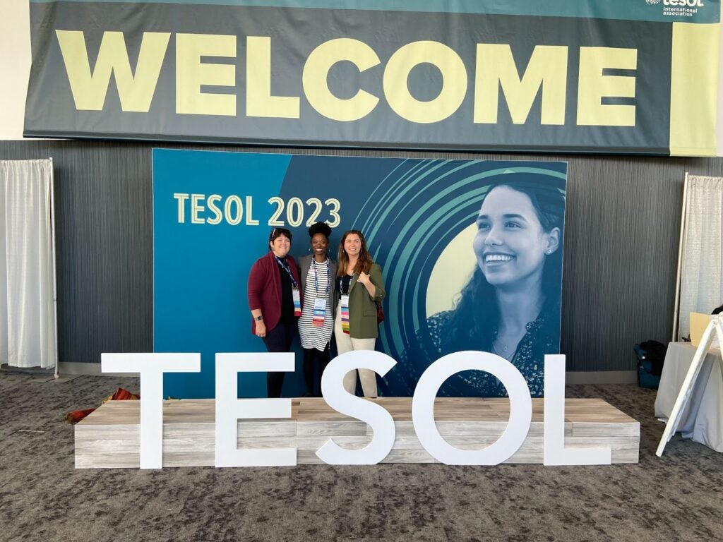 Katherine Anderson standing and posing with 2 other women in front of a large TESOL 2023 welcome sign inside a venue