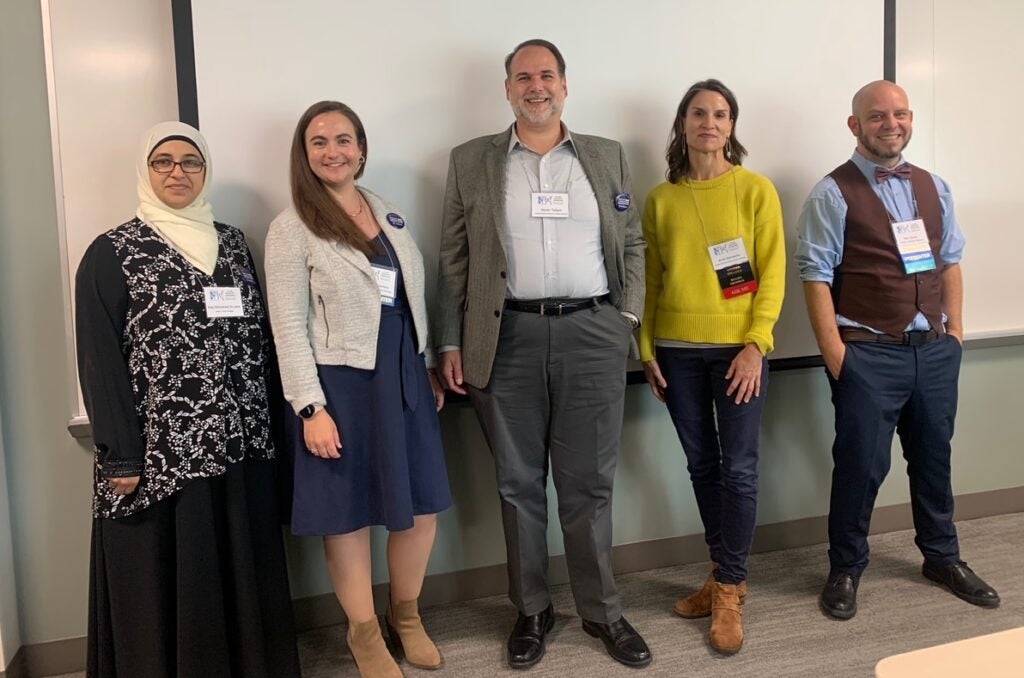 5 English Language Programs alumni, including Virtual Educator Anne Damiecka, pose for the camera standing in front of a projector screen that is just in front of a whiteboard