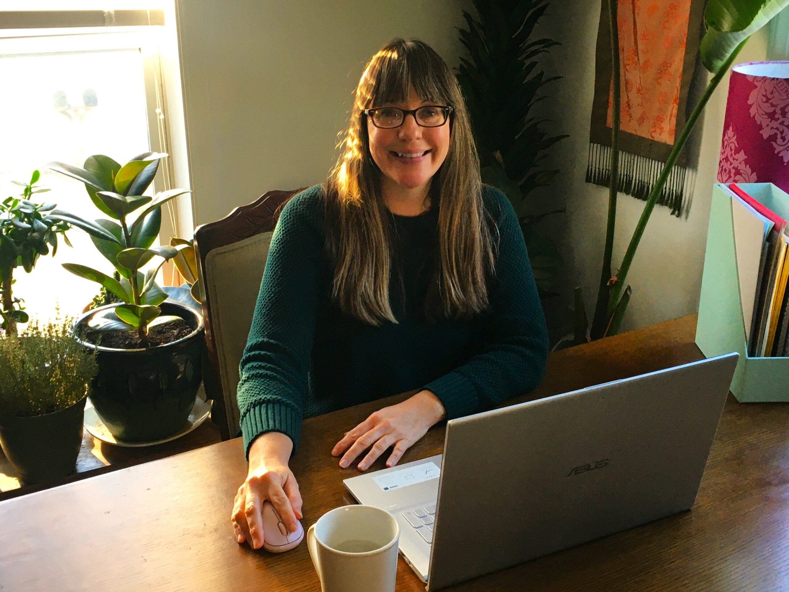 A Virtual Educator sitting behind her laptop with a mug at hand and plants in the background.